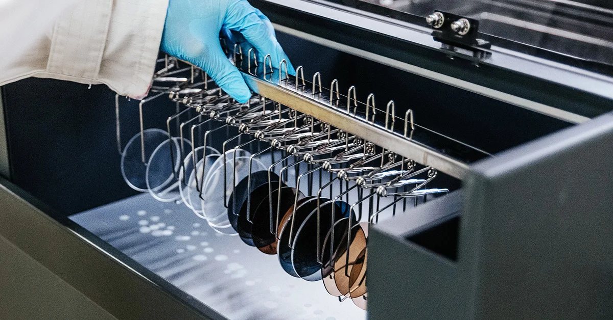 Mulher jovem de cabelo comprido e escuro, com óculos de armação preta, segura uma câmara fotográfica preta, apoiando o queixo nas mãos cruzadas sobre o equipamento. Veste uma camisa branca e usa um relógio metálico e uma pulseira preta. O fundo é em tons suaves de cinzento e lilás, criando uma atmosfera moderna e profissional. A imagem transmite confiança, criatividade e estilo, evocando o universo da fotografia e da precisão visual.